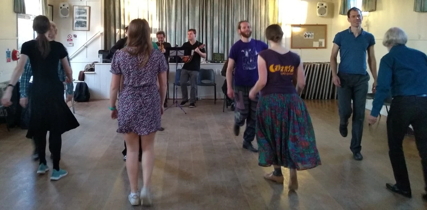 8 people dancing in a village hall. They are in 2 lines facing each other, accompanined by a trumpet and a mandolin.